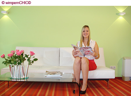 Das wimpernCHIC© Studio in Frankfurt ist im modernen wohnlichen Style eingerichtet. Die Wände sind im Pastell-Grün. In Oragne gehaltene Teppiche. Ein Glastisch mit frischen rosa-roten Rosen. Ein weißes weiches Sofa. Auf dem Sofa sitzt eine zufriedene Kundin. Sie blättert in einer Zeitschrift und lächelt dabei in die Kamera.